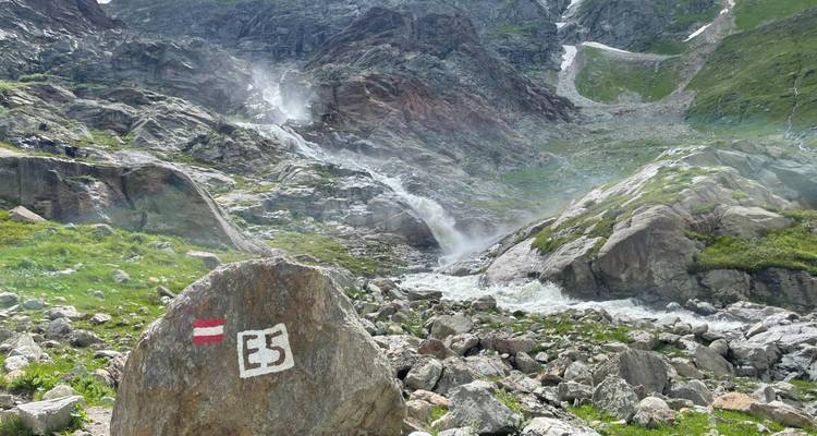 Rocher avec marqueur de sentier E5 à côté d'une cascade alpine en chute et d'un champ de rochers accidentés.