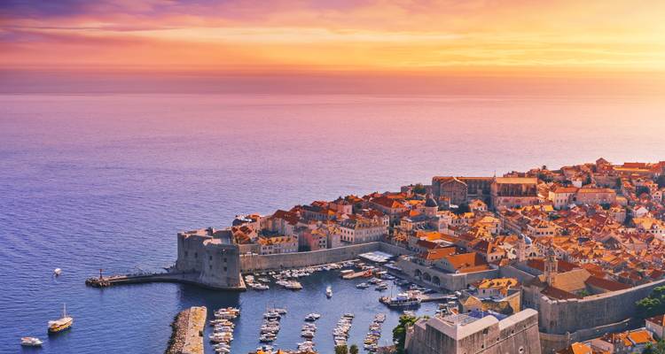 Coastal city with stone walls at sunset and a harbor full of boats.