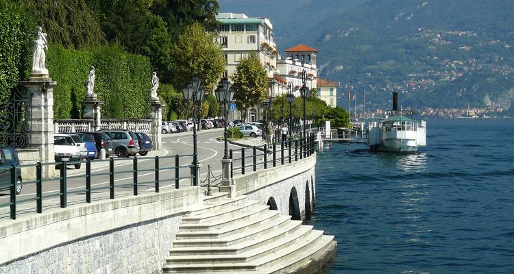 Une élégante route de promenade serpente le long du lac de Côme avec un bateau à vapeur à aubes historique amarré près des marches de pierre.