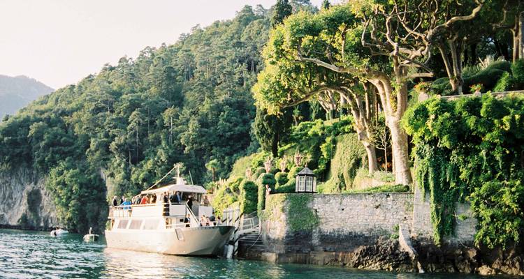 Bateau touristique amarré à côté des jardins en terrasses luxuriants d'une villa au bord du lac sous une douce lumière du soir.