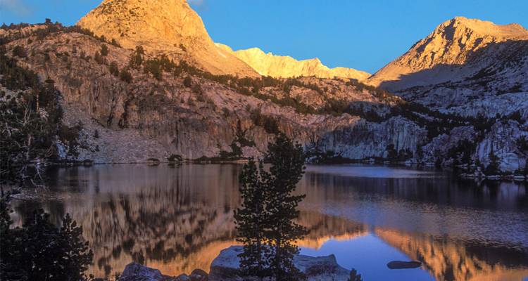 Un lago alpino refleja picos dorados al amanecer con pinos rodeando la orilla.