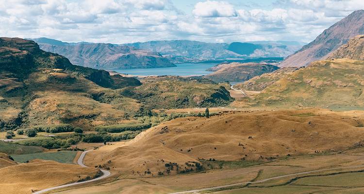 Vaste haute campagne néo-zélandaise aux collines dorées, routes sinueuses et lac turquoise lointain sous des nuages épars.