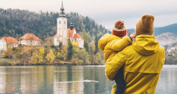 Un parent en manteau jaune tient un enfant tout en regardant à travers le lac de Bled vers l'église de l'île au milieu des couleurs d'automne.