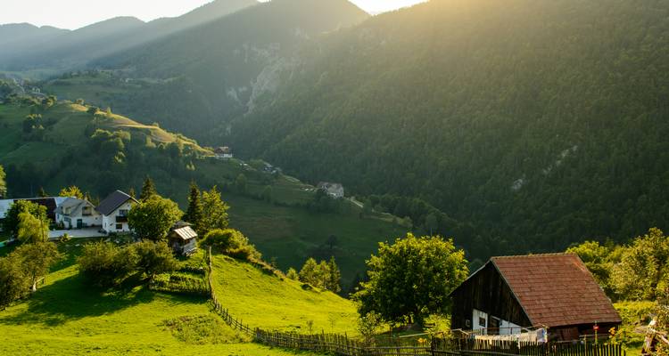 Sonnendurchflutetes ländliches Bauernhaus auf einem grünen Berghang, umgeben von steilen bewaldeten Tälern in der Dämmerung.