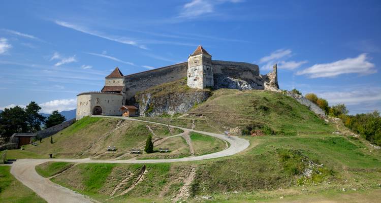 Mittelalterliche Festung Rasnov, die einen grasbewachsenen Hügel dominiert, mit einem gewundenen Weg unter blauem Himmel.