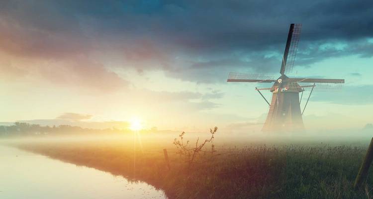 Un molino de viento holandés solitario emerge de la niebla del amanecer junto a un canal inmóvil mientras sale el sol.