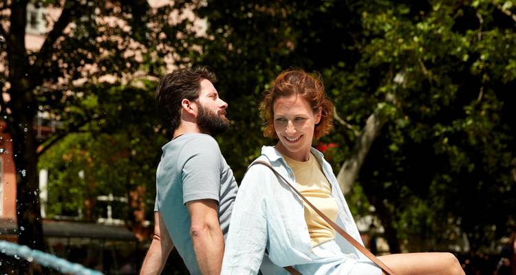 Jeune couple se détendant dos à dos près d'une fontaine urbaine ludique par une journée ensoleillée.