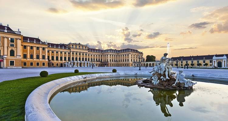 La larga fachada amarilla del Palacio de Schönbrunn y la estatua de la fuente se reflejan en un estanque tranquilo bajo cielos de atardecer pastel.