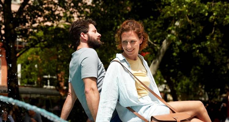 Una pareja sonriente se relaja espalda con espalda en el borde de una fuente en una plaza urbana frondosa.