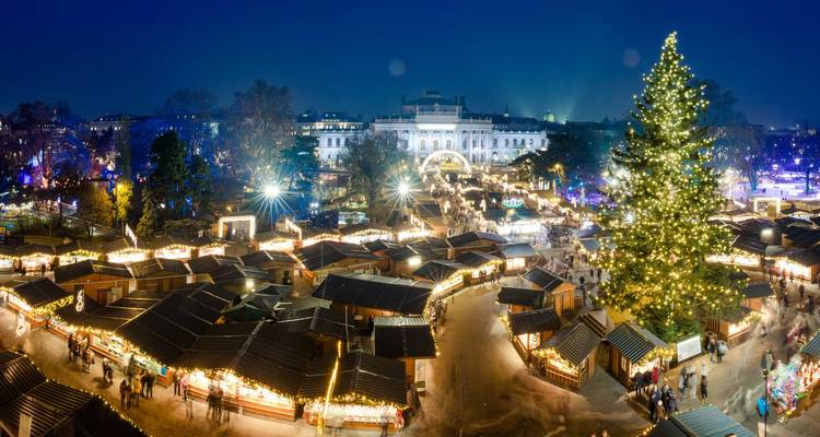 Vue aérienne nocturne du marché de Noël de Vienne avec ses petits chalets scintillants, sa patinoire et un immense sapin illuminé devant l'Hôtel de Ville.