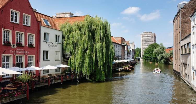 Canal verdoyant à Gand bordé de maisons rouges et blanches, de restaurants au bord de l'eau et d'un petit bateau de visite sous un ciel bleu.