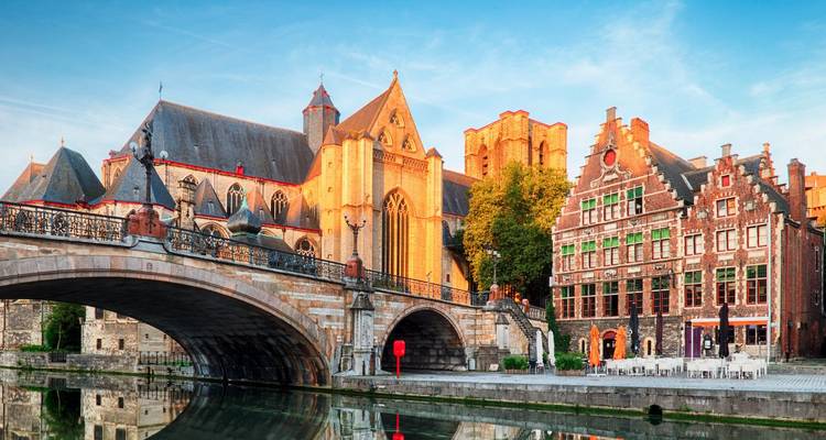 Scène de canal médiéval à Gand avec pont de pierre, églises historiques et lumière chaude du soleil sur les façades de briques
