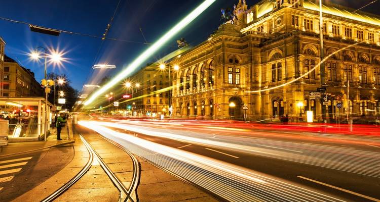 Scène nocturne en pose longue d'un grand opéra avec des traînées de lumière de tramway et un piéton solitaire.