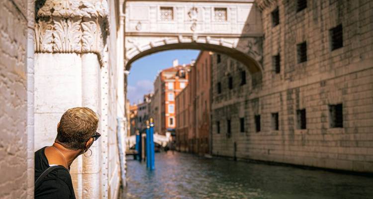 Ein Reisender blickt um eine Marmorecke zur Seufzerbrücke von Venedig, die sich über einen ruhigen Kanal wölbt.