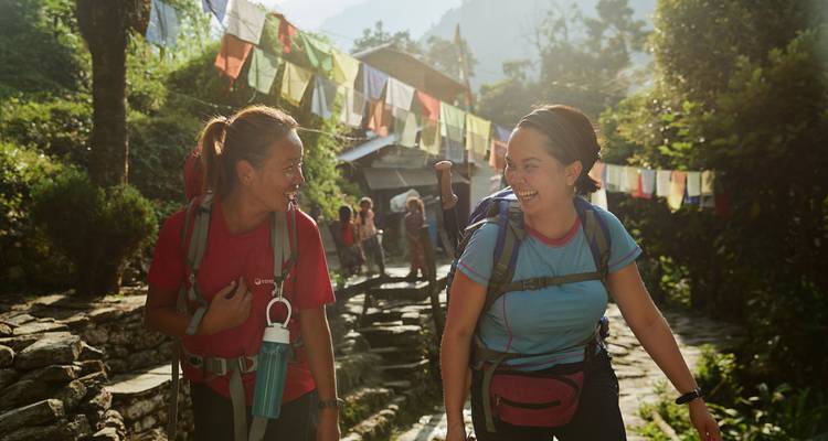 Two smiling trekkers walk beneath lines of colorful prayer flags on a lush Himalayan trail.