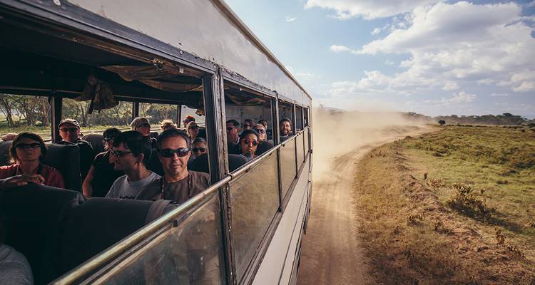 People seated in a bus looking out at a dirt road creating a cloud of dust.