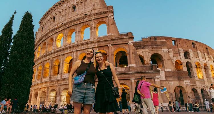 Deux voyageurs souriants posent devant le Colisée illuminé au crépuscule avec des foules qui se promènent alentour.