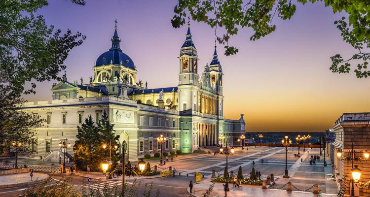 Vista al anochecer de la Catedral de la Almudena iluminada con farolas y ramas de árboles enmarcando la escena.