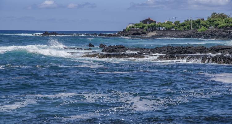 Les vagues s'écrasent sur des roches volcaniques sombres près d'un petit chalet côtier et d'arbres.