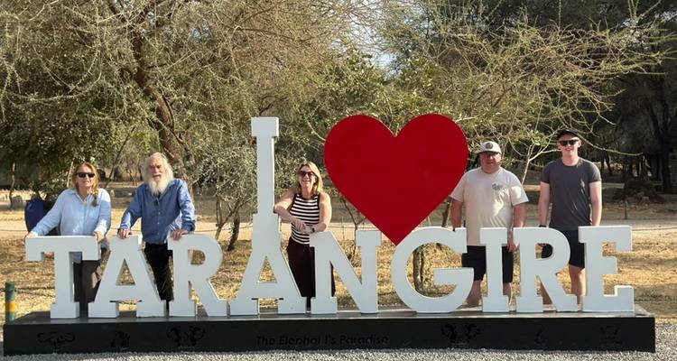 Cinco viajeros están de pie junto a un gran cartel que dice 'I ♥ Tarangire' en medio de un bosque seco.