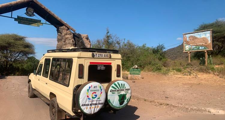 Un jeep de safari pasa por una entrada con un arco de piedra hacia un parque nacional bajo cielos azules.