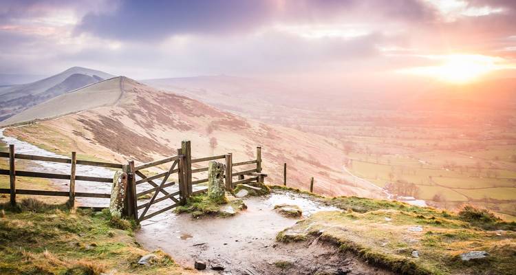 L'aube baigne les collines ondulantes du Peak District dans une lumière rose tandis qu'une porte en bois s'ouvre sur un sentier de crête.