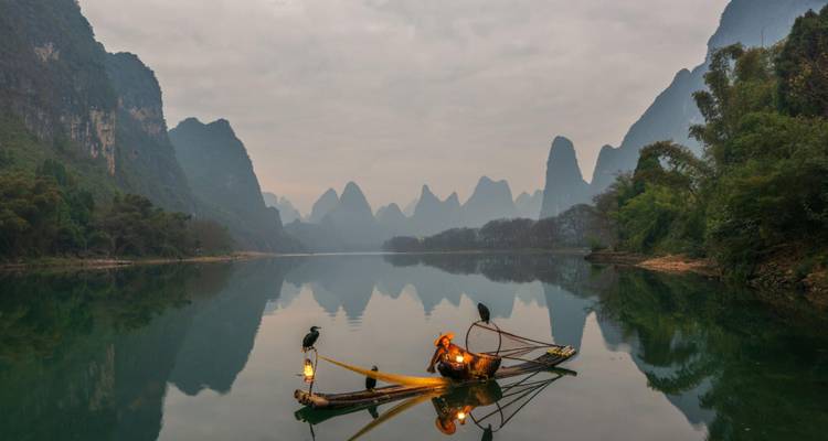Pescador tradicional de cormoranes en una balsa de bambú al amanecer entre picos de piedra caliza reflejados en aguas tranquilas.