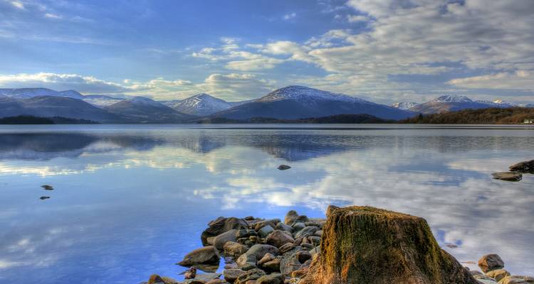 Lac serein avec premier plan rocheux, reflétant les sommets montagneux.