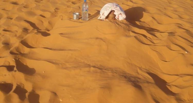 Personne partiellement ensevelie dans le sable avec une bouteille d'eau et une tasse sur un paysage désertique.