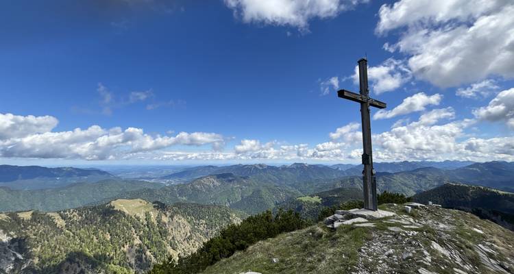 Cross at the peak with panoramic view of mountains and sky.