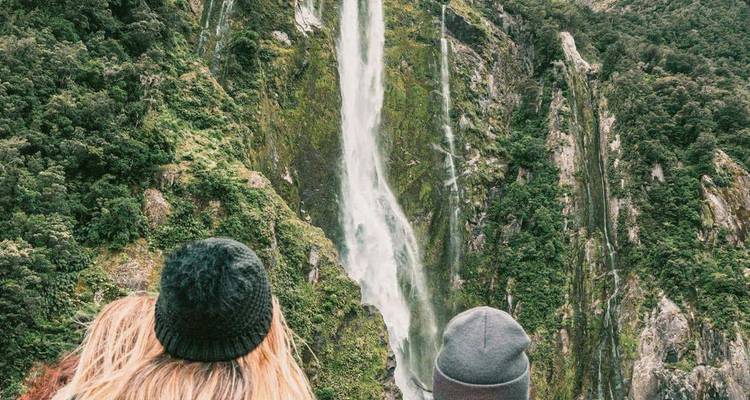 Two travellers in wool hats admire a towering rainforest waterfall cascading down a cliff