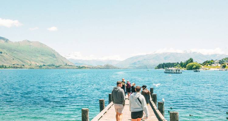 Des voyageurs marchent le long d'une jetée en bois au-dessus des eaux turquoise du lac Wanaka vers les paysages alpins environnants.