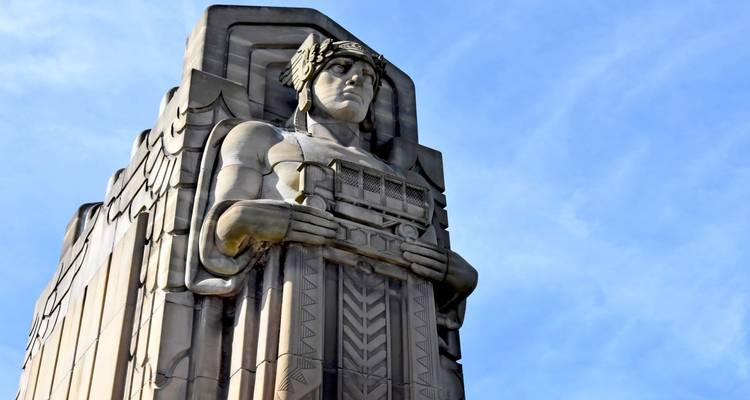 Gros plan d'une statue de gardien en pierre Art Déco tenant un panneau en relief contre un ciel bleu.
