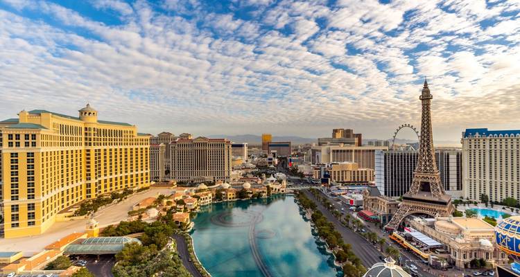 High vantage over the Las Vegas Strip showing Bellagio fountains, Paris Eiffel replica and casino skyline under patterned clouds.