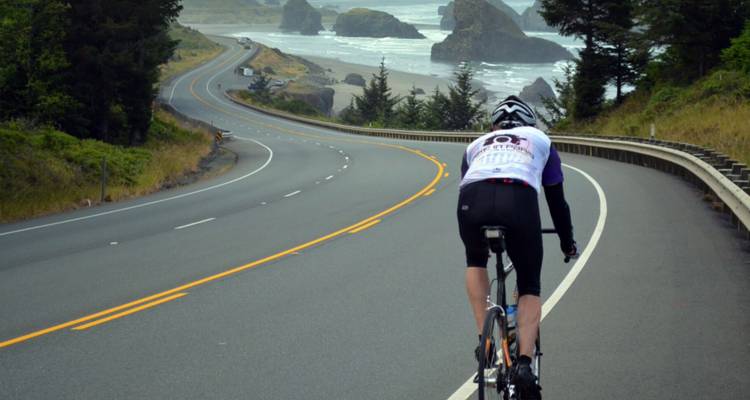 Cycliste sur une route côtière pittoresque avec des formations rocheuses et l'océan en arrière-plan.