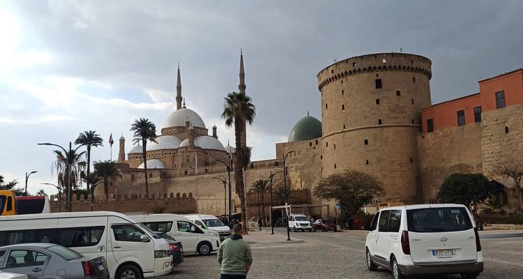 Personas y vehículos frente a una mezquita fortificada histórica bajo un cielo nublado.
