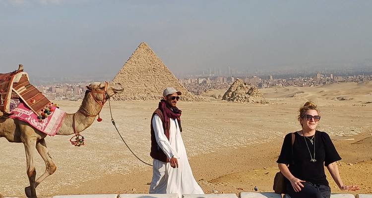 Turista y lugareño con un camello cerca de las pirámides, con vista al paisaje urbano.