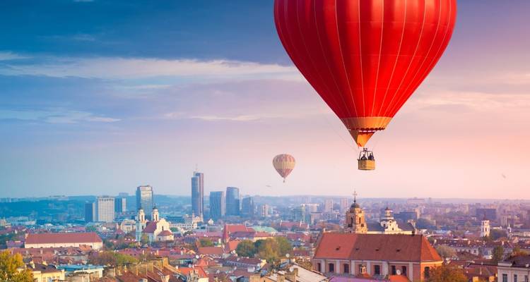 Hot-air balloons drifting over Vilnius old town roofscape during pastel sunrise.