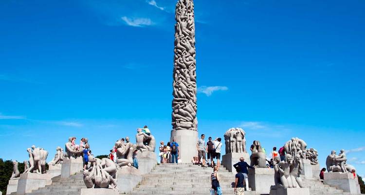 Los visitantes suben los escalones del Monolito de Vigeland en Oslo, un imponente pilar de granito de figuras entrelazadas bajo un cielo azul despejado.
