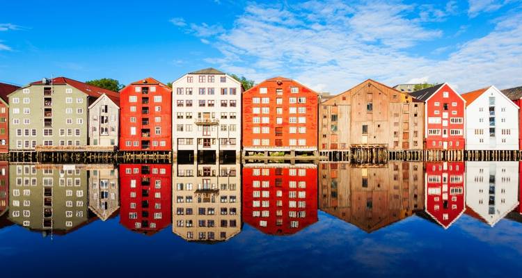 Almacenes tradicionales sobre pilotes de Trondheim reflejados perfectamente en las aguas tranquilas del río Nidelva bajo un cielo brillante.
