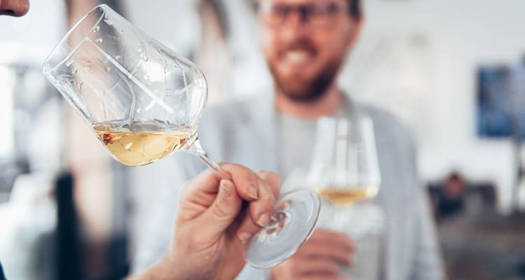 Personne tenant un verre de vin blanc avec une autre personne souriante en arrière-plan.