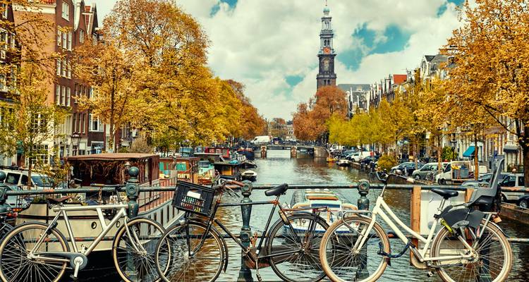 Amsterdamer Kanal mit Fahrrädern und Herbstbäumen, die einen historischen Turm einrahmen.