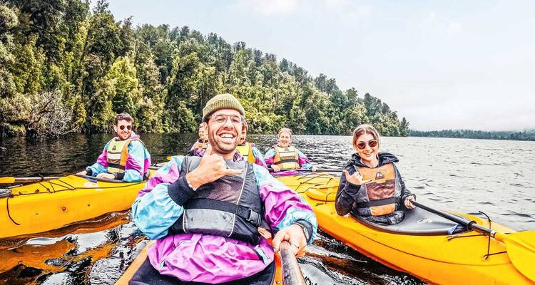 Kayakistas en botes amarillo brillante reman en un lago tranquilo bordeado de bosque, sonriendo a la cámara.