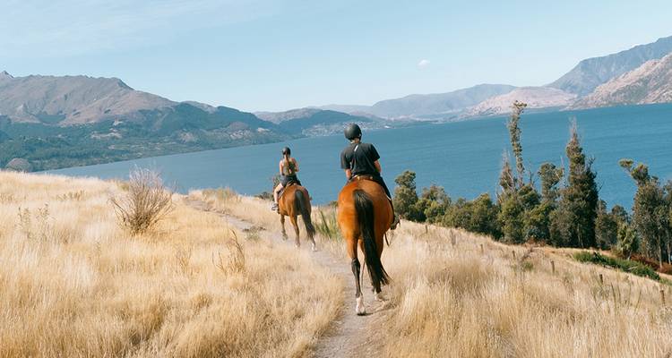 Dos jinetes a caballo siguen un sendero cubierto de hierba con vista a un lago alpino azul profundo y colinas distantes.