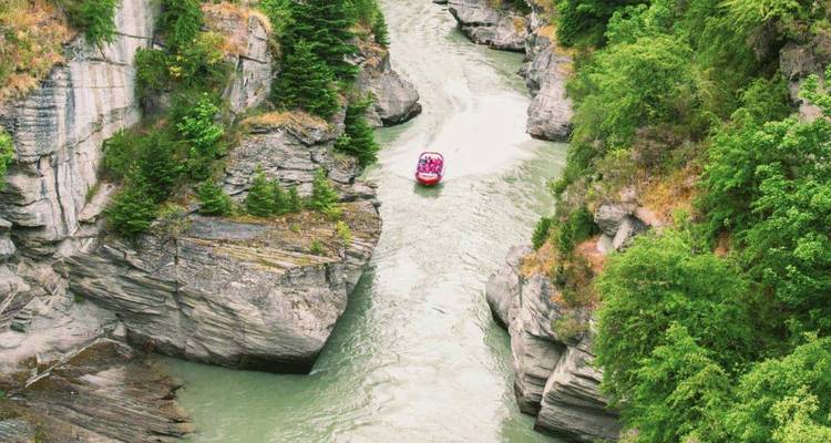 Red jet boat speeds through a narrow green river canyon flanked by tall rock walls and pines.