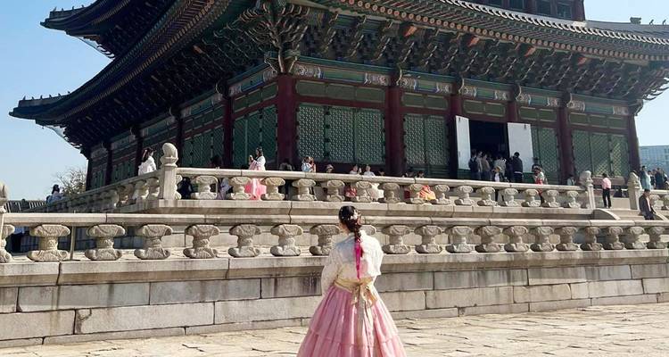 Sala tradicional del palacio coreano con visitantes; mujer en hanbok rosa de pie en el patio en primer plano.