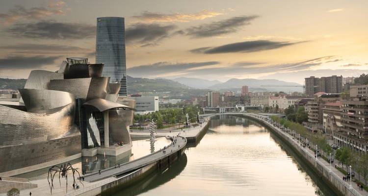 La luz del amanecer baña el Museo Guggenheim Bilbao junto al río reflejante y las colinas distantes.