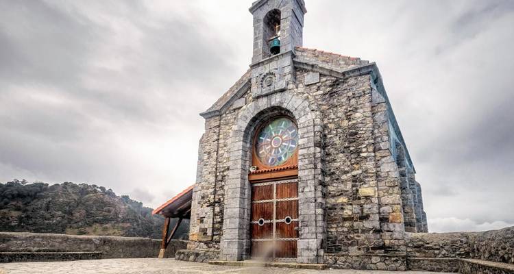 Capilla de piedra con rosetón de vitrales situada en una cresta montañosa brumosa bajo cielos nublados