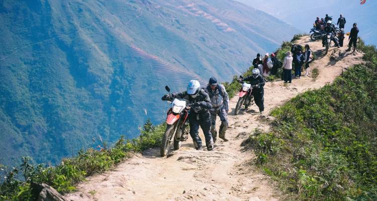 People pushing motorcycles on a beaten path with a hilly backdrop.