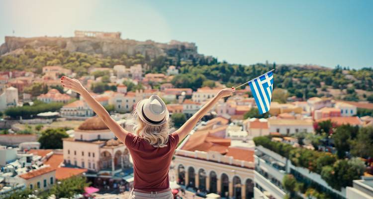 Frau mit ausgebreiteten Armen, die über Athen blickt, mit einer griechischen Flagge.